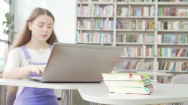 Kendini işine adamış genç bir kız, parlak bir kütüphanede canlı kitaplarla çevrili dizüstü bilgisayarında dikkatlice yazıyor. Bu sahne akademik arayışın özünü yakalıyor, öğrenme ve keşfe olan bağlılığını gösteriyor.