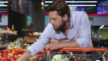 Havalı sakallı, modaya uygun genç bir beyaz adam, hareketli bir süpermarkette olgun domates ve salatalıkları değerlendiriyor. Onun keskin gözleri ve düşünceli ifadesi canlı renkler ve cazip gösteriler gibi taze, sağlıklı ürünlere olan tutkusunu yansıtıyor.