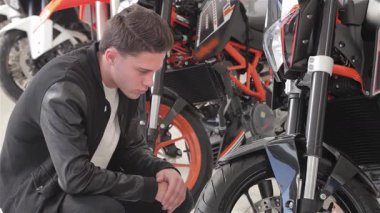 Neşeli beyaz bir adam, çarpan siyah bir motosikleti yakından incelerken onay veriyor. Motosikletlerin parlak tasarımlarına ve güçlü özelliklerine hayran olduğu için heyecanı yayılıyor.