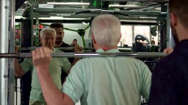 Kararlı bir beyaz adam spor salonunda antrenman yapıyor, omuzlarında bir halterle çömeliyor. Güçlü bir antrenman yaparken atletik ruhu parlıyor. Destekleyici bir antrenör aynadaki yansımasını yakından izliyor.