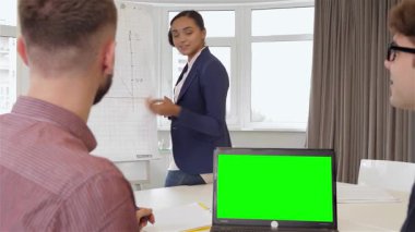 Dengeli bir siyahi kadın projesini parlak ve modern bir ofisteki iki erkek meslektaşına tutkuyla sunar. Bir grafiğin yanında durup dikkatlerini çekerken yeşil renkli ekranlı bir dizüstü bilgisayar masanın üzerinde duruyor.