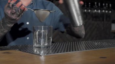 Dövmeli bir erkek barmen tarafından ustalıkla bardağa dökülen sanatsal bir kokteyl miksoloji yeteneklerini sergiliyor. Barın hareketli atmosferi, retro dekoru ve yumuşak ışıklandırmasıyla, gece hayatının cazibesini ve el yapımı zevkini arttırır.