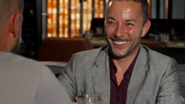 Gri takım elbiseli neşeli, olgun bir adam sofistike bir viski barında arkadaşıyla gülüp sohbet ediyor. Sıcak ortam ve zarif dekor, onların neşeli etkileşimlerini güçlendiriyor, arkadaşlığı ve eğlenceyi coşkulu bir şekilde kutluyorlar.