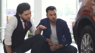 İki şık genç adam modern bir galeride lüks bir otomobilin tekerlek ve lastiklerini inceliyor. Araçların özelliklerini tartışırken yoldaşlıkları parlıyor, arkadaşlık ve macera dolu bir yaşam tarzını somutlaştırıyor.
