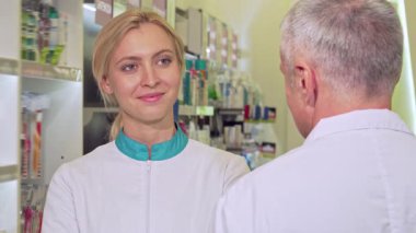 Neşeli bir kadın kimyager, kıdemli meslektaşıyla dolu bir eczanede canlı bir sohbet ederken neşeyle ışıldıyor. Parlak, davetkar atmosferde renkli ilaç ürünleriyle dolu raflar sergileniyor.