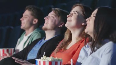 Çok katlı bir tiyatroda oturan dört arkadaş, ekrandaki film karşısında büyülendiler, yüzleri heyecan ve neşe içinde parlıyordu. Elinde patlamış mısırla ve her yerde gülümsemeyle bu canlı sahne, arkadaşlığın ve sinemanın heyecanını yakalar.