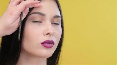 Yetenekli bir vizojist güzel bir Asyalı kadının saçını incelikle şekillendirir, göz makyajına hazırlanırken yüzünü güzelleştirir. Parlak sarı arka plan canlı bir dokunuş ekler, esmer modellerin zarafetini vurgular.