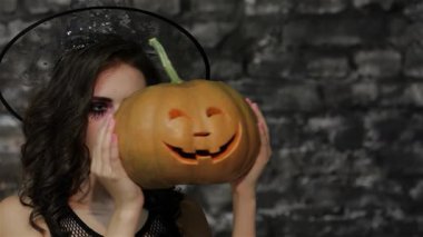 Büyüleyici bir kadın-büyücü yüzünü dramatik bir siyah tuğla duvara dayamış sırıtan bir balkabağının arkasına saklar. Bu Cadılar Bayramı temalı görüntü gizem ve sihir yayarak izleyicileri büyüler dünyasına davet ediyor.