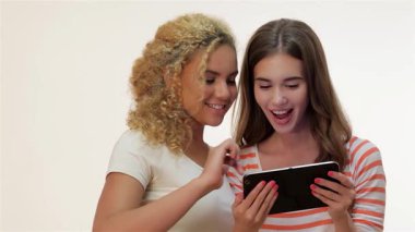 Birinin kıvırcık sarı saçlı, diğerinin uzun kahverengi saçlı iki kadın, yumuşak beyaz bir arkaplan üzerinde bir tablet bilgisayarla heyecan verici bir şekilde etkileşime geçiyorlar. Neşeli ifadeleri ve günlük kıyafetleri neşe dolu bir atmosfer yaratıyor.