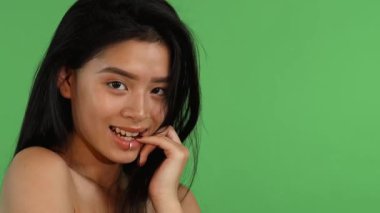 Büyüleyici genç Asyalı bir kadın canlı yeşil renkli bir arka plana karşı poz veriyor. Parlak bir gülümsemeyle parmağını ısırıyor. Doğal güzelliği ve kendine güveni, neşe dolu davetkar bir atmosfer yaratıyor.