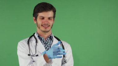 Çekici sakallı bir erkek doktor mavi lastik eldivenlerini giymeye hazırlanırken kendine güvenle gülümsüyor, parlak yeşil renkli bir arka plana karşı. Bu ilgi çekici sahne, tıp alanındaki profesyonelliğin ve tedavinin özünü yansıtıyor.