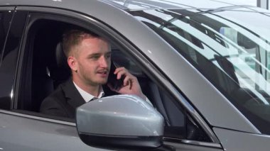 Kendine güvenen bir işadamı, şık bir takım elbise giymiş, modern otomobilinin rahatlığıyla bir telefon görüşmesi yaparken gülümsüyor. Işıldayan iç mekan, lüks ve teknolojinin bir karışımını yansıtıyor, yeni mülkiyetin heyecanını ve