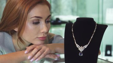 Kuyumcuda sergilenen zarif elmas kolyeye özlemle bakan güzel, üzgün bir kadının yakın çekimi. İfadesi hayal kırıklığını ve hasreti yansıtıyor. İhtirasın duygusal ağırlığına karşı göz kamaştırıcı bir maliyet.