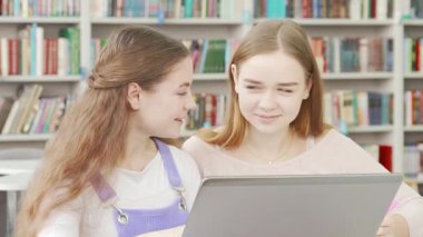 İki neşeli genç kız kitaplarla dolu renkli bir kütüphanede dizüstü bilgisayarlarını paylaşıyorlar. Ödevlerini yaparken arkadaşlıkları parlıyor, öğrenme ve iletişim ruhunu somutlaştırıyor.