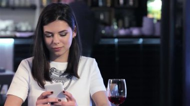 Şık bir esmer kadın modern bir restoranda oturmuş elinde bir bardak zengin kırmızı şarap tutarken dikkatle akıllı telefonuna bakıyor. Ortam sofistike, hafif ışıklandırma onun rahatlamış tavrını arttırıyor ve şaraptan bir yudum alıyor, mükemmel bir şekilde.