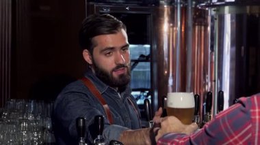 Çekici sakallı bir barmen, müşterisiyle meşgul olurken zengin bir koyu biranın tadını çıkarır. O da kadehini köpüklü bardakla kaldırır. Bu canlı bar sahnesi yoldaşlığın ve kutlamanın özünü yakalıyor. Oktoberfest havası için mükemmel.