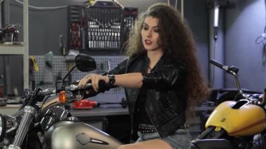 Heyecanlı, kıvırcık saçlı, siyah deri ceketli genç bir kadın, hareketli bir atölyede motosikletinin üzerinde otururken başparmağını kaldırıyor. Bu çekici beyaz motorcu neşe ve güven saçıyor, heyecanı somutlaştırıyor.