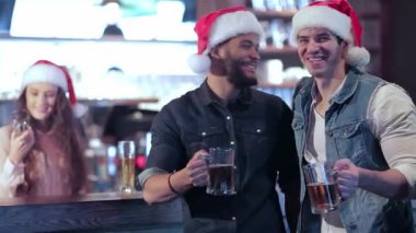 Noel Baba şapkalı iki arkadaşın bira bardaklarını kaldırıp kalabalık bir barda tatil ruhunu kutladıkları şenlikli bir toplantı başlıyor. Neşeli barmen arka planda gülümsüyor, kahkaha ve arkadaşlıkla dolu coşkulu atmosferi de ekliyor.