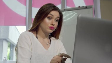 Uzun saçlı neşeli genç bir kadın dizüstü bilgisayarında canlı üniversite kütüphanesi atmosferiyle çevrili bir görüntülü sohbete dalarken ışıl ışıl gülümsüyor. Ailesiyle bağ kurarken, modern eğitimin ruhunu somutlaştırırken coşkusu yayılıyor.