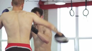 Güçlü bir kickboksçunun koçuyla güçlü yumruklar atması, azim ve beceri göstermesi gibi yoğun bir sahne gözler önüne seriliyor. Eğitim ekipmanlarıyla dolu parlak spor salonu, bu dinamik çalışmanın enerjisini arttırıyor,