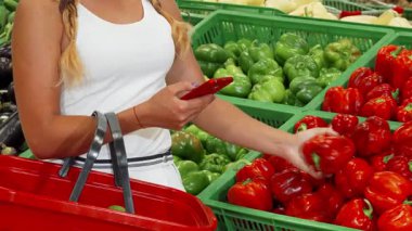 Beyaz elbiseli şık bir kadın, süpermarkette taze sebze denizinin ortasında canlı kırmızı biberleri seçerken akıllı telefonuyla meşgul oluyor. Bu canlı sahne modern alışverişin özünü yakalıyor, teknolojiyi zevkle harmanlıyor.