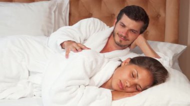 Çekici bir çift, lüks bir otel yatağında, yumuşak bornozlara sarılmış, sükunet ve sevgi dolu bir şekilde uzanıyor. Yakışıklı genç adam karılarını nazikçe okşar. Birlikte huzurlu bir anın tadını çıkarırlar ve samimi bir ortam yaratırlar.