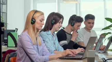 Kulaklık takan odaklanmış genç bir kadın kendini işine adamış meslektaşlarıyla dolu canlı bir çağrı merkezinde çağrılara cevap veriyor. Parlak, modern çalışma alanı yeşil ve teknolojiyle dolu, takım çalışması için enerjik bir atmosfer yaratıyor.