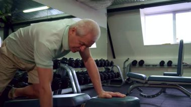 Yaşlı, beyaz bir adam, spor salonunda dizini dinlendirirken halterini kaldıran odaklanmış bir çalışmayla meşgul. Genç bir antrenör yanında duruyor, destek ve cesaret veriyor, bu fitness limanında motive edici bir atmosfer yaratıyor.