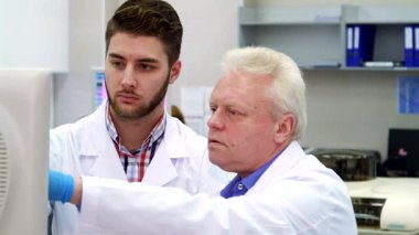 Modern bir laboratuvarda, üst düzey bir Kafkas bilim adamı odaklanmış genç bir stajyere rehberlik ederek bir monitörü tutkuyla işaret ediyor. Yakın çekim onların yoğun işbirliğini yakalıyor, canlı bir bilimsel ortamda bilgi alışverişini gösteriyor.