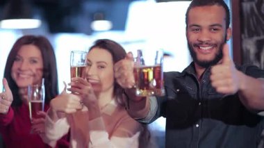 Aralarında iki kadın ve bir erkeğin de bulunduğu genç bir grup, coşkulu bir şekilde yükseltilmiş bira bardaklarıyla tezahürat yapıyor, hareketli bir barda arkadaşlığı kutluyorlar. Parlak gülüşleri ve baş parmakları neşe saçıyor, arkadaşlığın özünü yakalıyor.