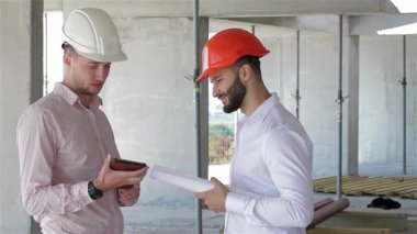 Biri beyaz şapkalı, diğeri turuncu şapkalı iki genç müteahhit, hareketli bir inşaat sahasında dijital tablette sergilenen planları canlı bir şekilde tartışıyor. Sakallı adam bir tomar plan tutarken, meslektaşı da...