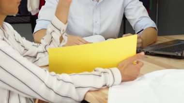 İki yetenekli genç bayan moda tasarımcısı, arka planda asılı renkli giysilerle çevrili canlı bir tartışmaya girişiyor. Yaratıcı sinerjileri ve takım çalışmaları, işbirliğinin ruhunu somutlaştıran yenilikçi fikirlerin beyin fırtınası gibi parlıyor.