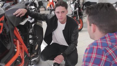 Bilge bir satıcı meraklı genç bir adama çarpan turuncu spor bir motosiklet sunar. Müşteri motosikletlerin düzgün çerçevesini ve rahat koltuğunu incelerken, danışman da onun etkileyici özelliklerini tutkuyla vurgular.