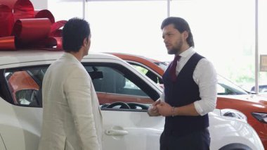 Sivri takım elbiseli sofistike bir araba satıcısı, kırmızı bir fiyonk ile süslenmiş gösterişli beyaz bir arabanın kapısını açar ve erkek müşterisini iç mekanı keşfetmeye davet eder. Araba galerisindeki bu heyecan verici an,