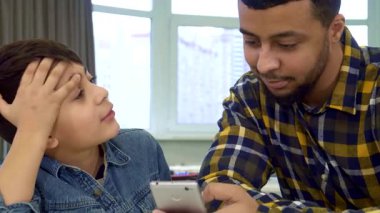 Afrika kökenli Amerikalı bir baba ve küçük oğlu çok hoş bir anda bir akıllı telefon ekranını yakından inceliyorlar. Küçük çocuk, düşünceli bir yüz ifadesiyle, babası ekose gömlek giyerken elini alnına koyar.