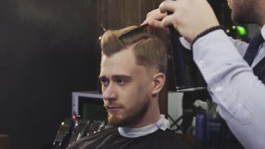 Odaklanmış genç bir adam berber koltuğunda güven içinde otururken profesyonel bir berber saçını saç kurutma makinesi ve saç spreyi kullanarak ustaca şekillendiriyor. Sahne, modern erkeksiliğin ve tımar etmenin özünü yakalıyor. Sanatçılık ve hassasiyeti gözler önüne seriyor.