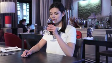 Güzel esmer bir kadın şık bir restoranda tek başına oturmuş, düşünceli bir ifadeyle kırmızı şarap yudumluyor. Uzun saçları, düşüncelere dalmış, düşüncelere dalmış, yalnızlık ve özlem hissi uyandıran bir yüzü çerçeveler.