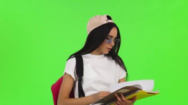 Neşeli genç bir bayan öğrenci, şık bir beyzbol şapkası ve şık güneş gözlükleriyle, renkli kitaplarını canlı yeşil renkli bir arka plana karşı tutarken pozitiflik saçıyor. Bu canlı sahne gençliğin ve eğitimin özünü yansıtıyor.