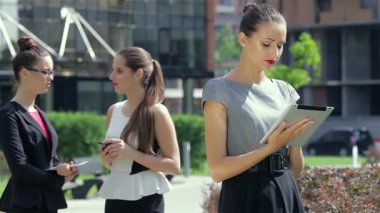 Kendine güvenen genç bir iş kadını dışarıda duruyor ve meslektaşları yakınlarda sohbet ederken dikkatle tabletini hazırlıyor. Bu dinamik sahne, modern iş işbirliğinin özünü yakalar.