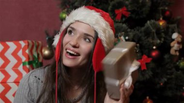 Çekici esmer bir kadın, Noel Baba şapkası takmış, güzel bir Noel hediyesi kutusunu sallıyor, yüzü heyecanla parlıyor. Göz kamaştırıcı bir Noel ağacıyla çevrili renkli süsler ve parıldayan ışıklarla süslenmiş,