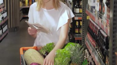 Sarı saçlı odaklanmış genç bir kız alışveriş listesini, hareketli bir süpermarket koridorunda dikilirken inceliyor. Renkli ürünlerle çevrili halde, taze sebzeleri ve sebzeleri dikkatli bir şekilde sınıflandırıyor, dikkatli olmanın özünü somutlaştırıyor.