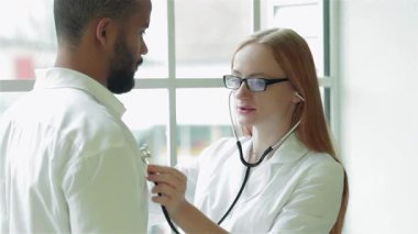 Parlak ve modern bir klinikte, odaklanmış gözlüklü kadın bir doktor erkek hastalarının kalp atışlarını stetoskop kullanarak nazikçe dinler. Sıcak atmosfer profesyonel etkileşimlerini vurguluyor. Reçete yazmaya hazırlanıyor.