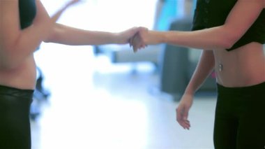 Hareketli bir spor salonunda, yakın plan çekimde bir kız el sıkışıyor, durağan bisikletlerle pedal çeviren arkadaşların arasında arkadaşlığı sembolize ediyor. Bu dinamik sahne enerji ve motivasyon yayar, takım çalışmasının ve formda olmanın ruhunu gösterir.