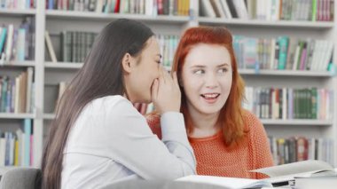 İki neşeli üniversiteli kız arkadaş renkli kitaplarla dolu, canlı bir kütüphanede fısıldayıp gülüyorlar. Canlı dostlukları ve sırlarını paylaşmaları dostluk ve işbirliği için davetkar bir ortam yaratıyor.