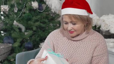 Noel Baba şapkası takan neşeli, yaşlı bir kadın kameraya doğru güzelce paketlenmiş hediyesini uzatıyor. Bu iç açıcı sahne, Noel neşesinin özünü yakalıyor, aile kutlamalarının sıcaklığını sergiliyor.