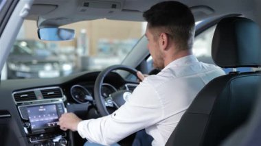 Sakallı neşeli bir beyaz adam galerideki gösterişli bir araba konsolunun dokunmatik ekranına zevkle dokunuyor. Aracın modern özelliklerini keşfederken, teknolojinin mükemmel bir karışımını sergileyerek, başparmak hareketi heyecan saçıyor.