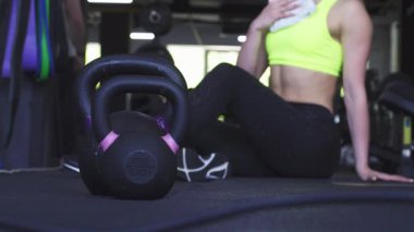 Spor salonunun zemininde kettlebell 'in odaklanmış bir görüntüsü yatarken, neon ışıklı tepeli atletik bir kadın elinde havluyla duvara yaslanıyor. Bu dinamik sahne kararlılığın ve gücün özünü yakalar, sert ve sert arasındaki dengeyi gösterir.