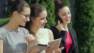 Üç kendine güvenen iş kadını bir ofisin dışında durmuş tabletlerini gözden geçirirken hararetli bir şekilde tartışıyorlar. Parlak gülümsemeleri ve şık kıyafetleri modern şehir ortamında canlı bir başarı ve takım çalışması atmosferini yansıtıyor.