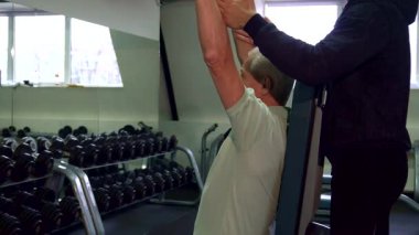 Kararlı bir Kafkasyalı son sınıf öğrencisi, spor salonunda omuz eğitimiyle meşgul, bir dambılı titizlikle kaldırıyor. Genç, sakallı bir antrenör onu arkadan destekliyor. Bu canlı fitness ortamında formda ve motivasyonlu olmasını sağlıyor.