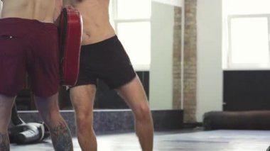 Dinamik bir vuruş profesyonel bir kickboksörü modern bir spor salonunda koçuyla güçlü tekmeler atarken yakalar. Kaslı vücudunu sergileyen atlet, tekmeye vururken tekniğini mükemmelleştirmeye odaklanıyor.